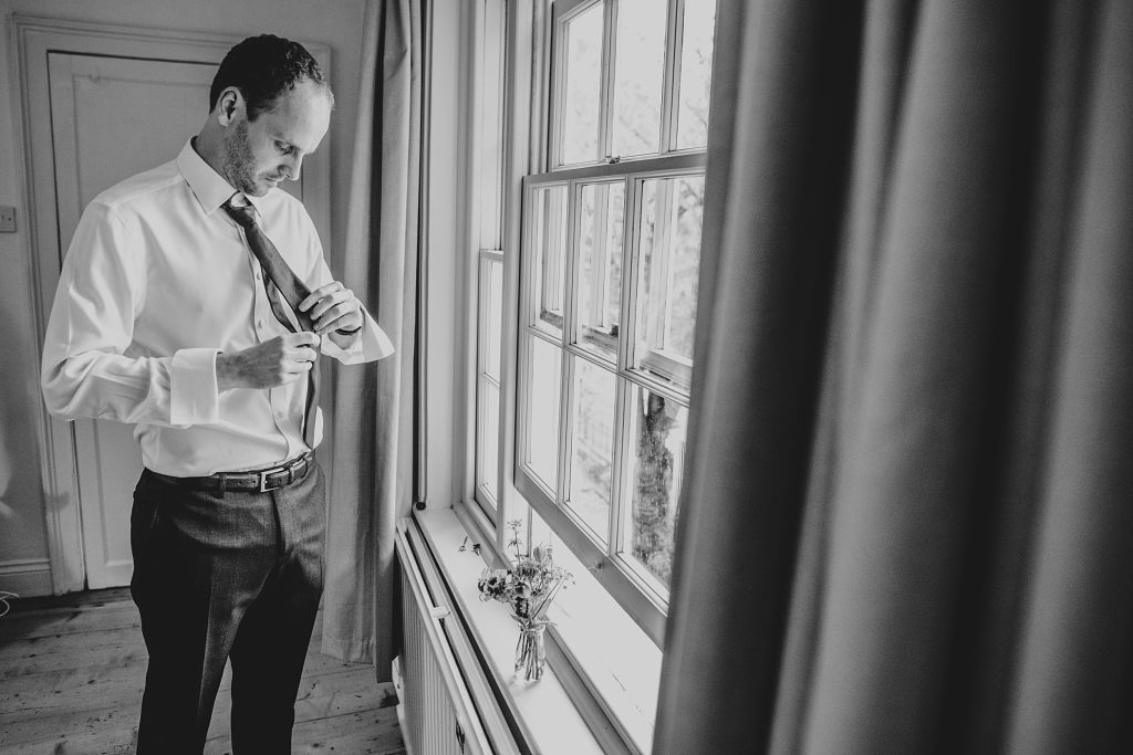 groom puts his tie on next to a window