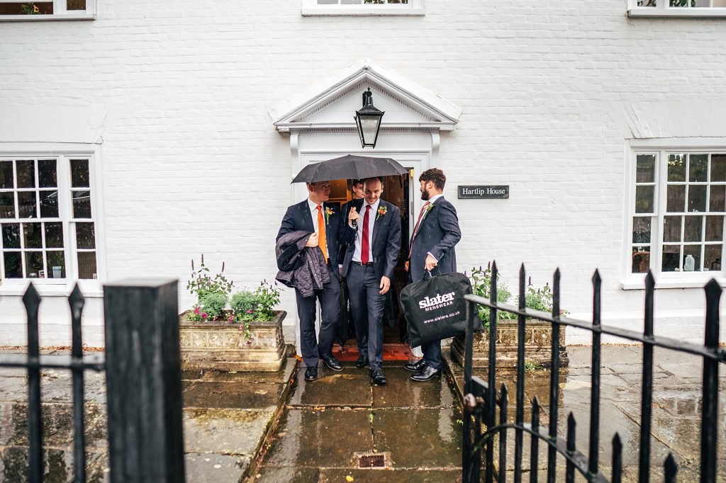 groom and groomsmen leave prep house under an umbrella in the rain