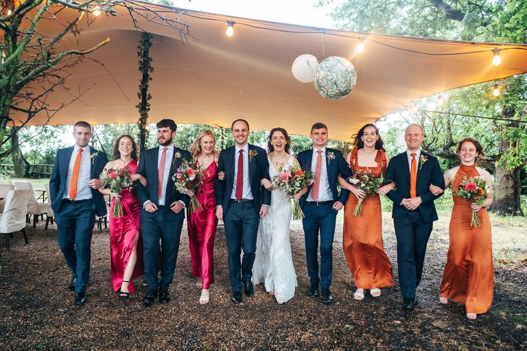 bride, groom and wedding party in pink and orange, link arms and walk towards camera under stretch canopy