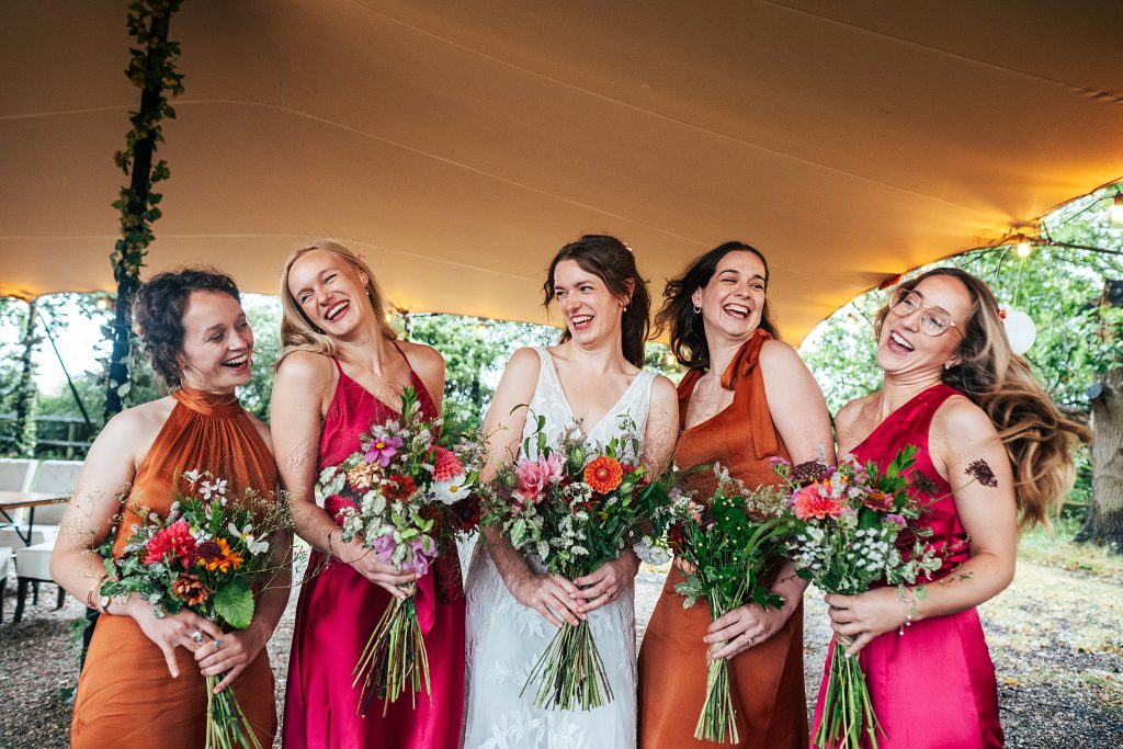laughing bride and bridesmaids in pink and orange stand under stretch canopy