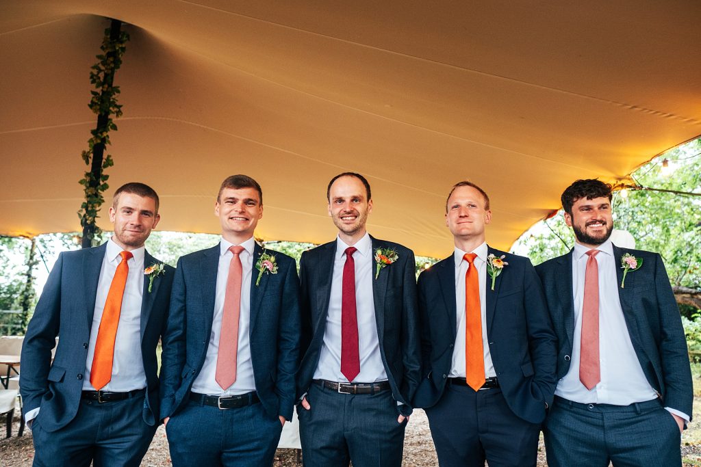 groom with groomsmen in navy in pink and orange ties stand under stretch canopy