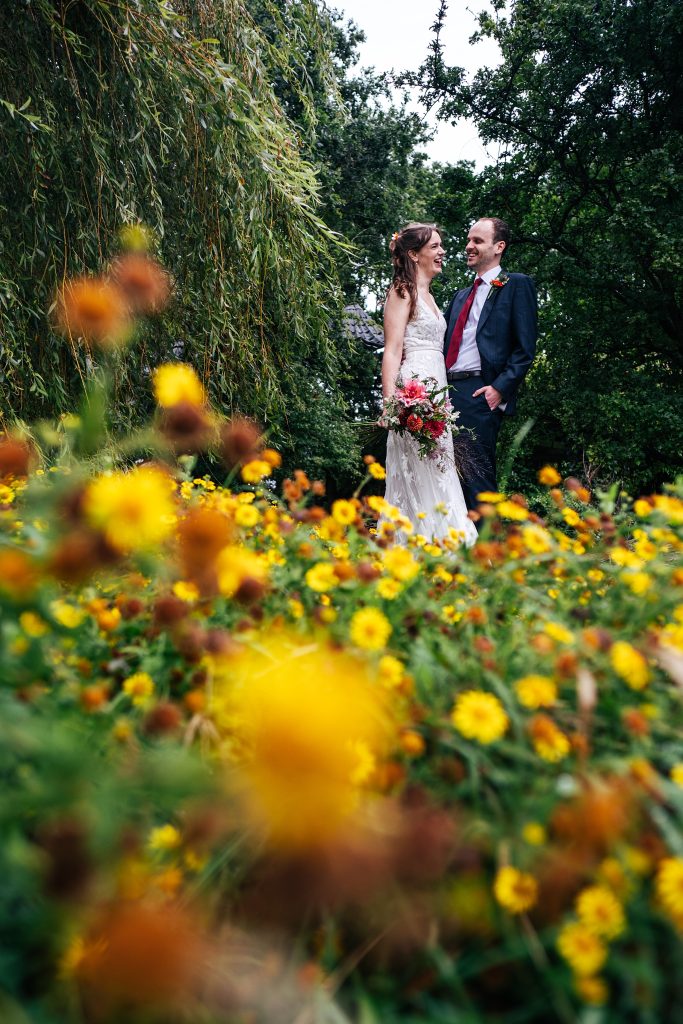 laughing bride and groom stand in a garden with yellow flowers in foreground