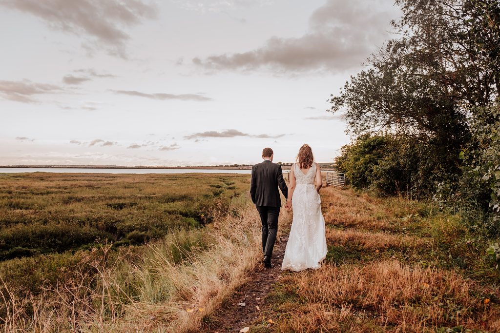 bride and groom hold hands and walk away from camera along a path to the coast at sunset