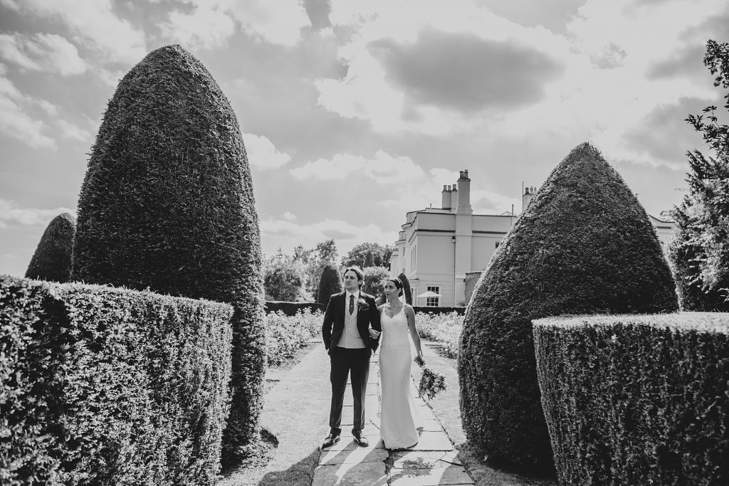 bride and groom stand between two manicured bushes in garden