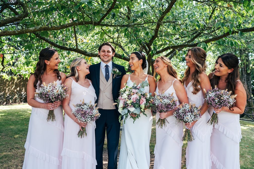 bride and bridesmaids all look at laughing groom under tree