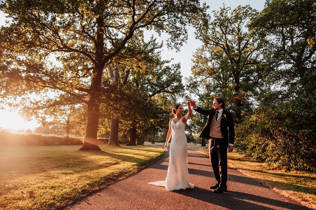 groom spins bride on treelined driveway at sunset