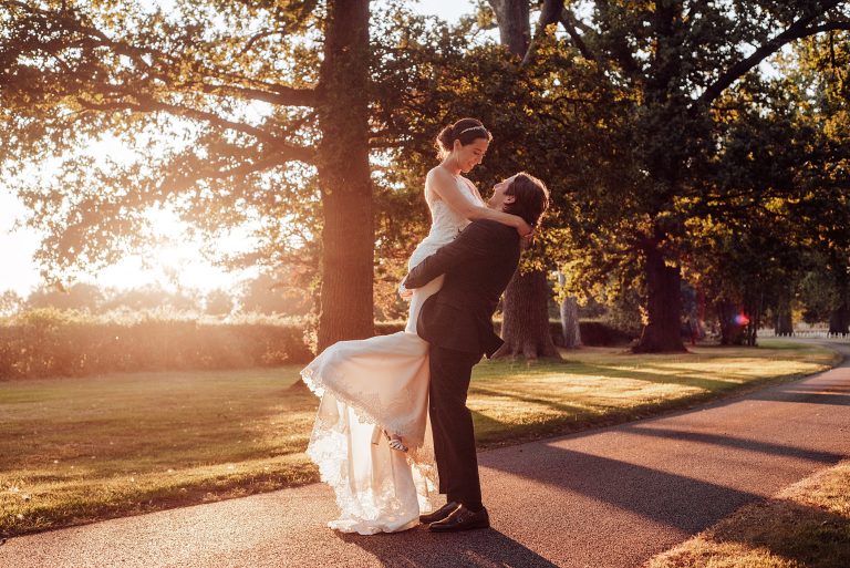 groom lifts bride on treelined driveway at sunset