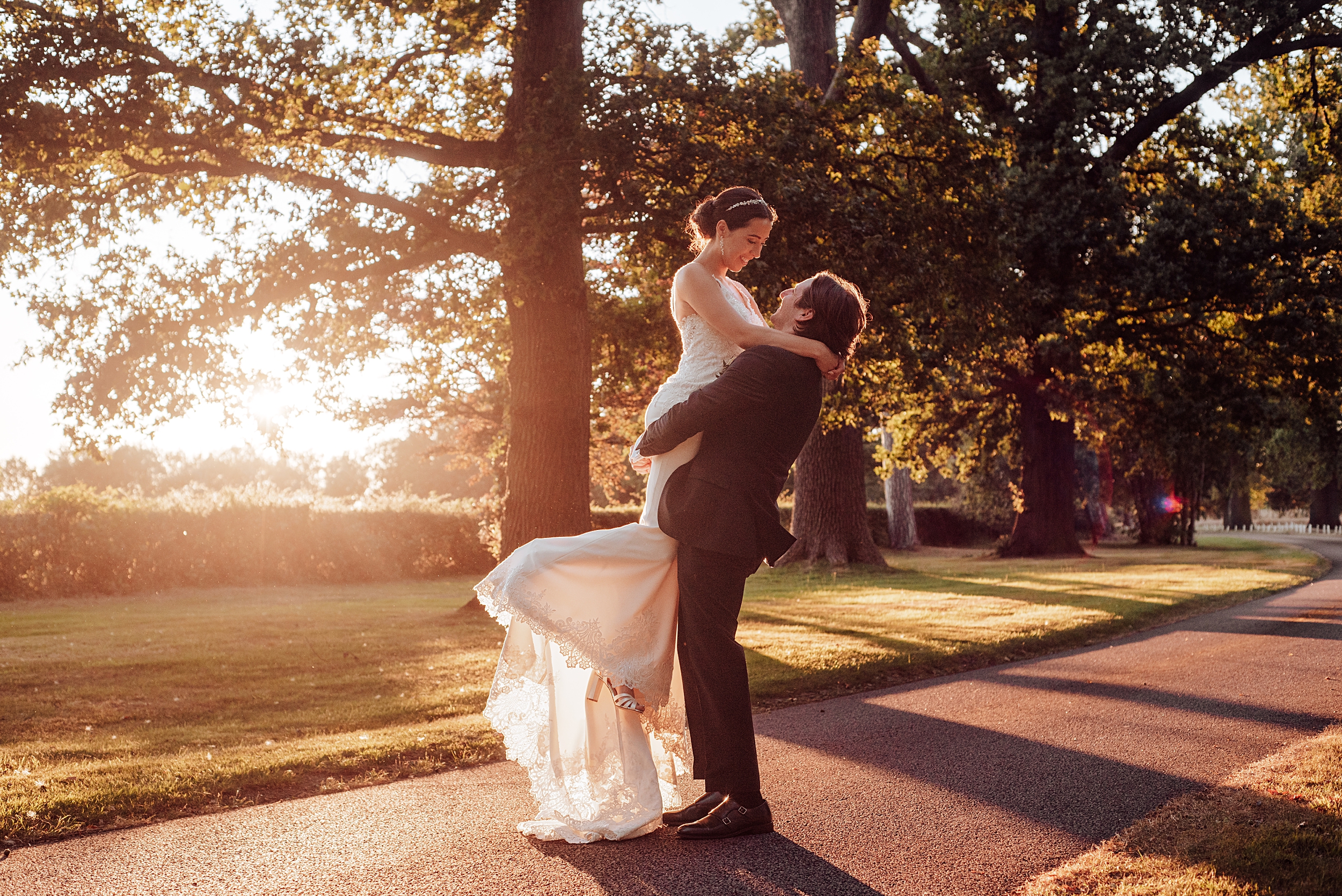 groom lifts bride on treelined driveway at sunset