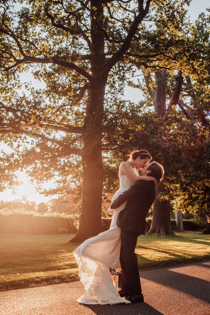 groom lifts bride and kisses her on treelined driveway at sunset