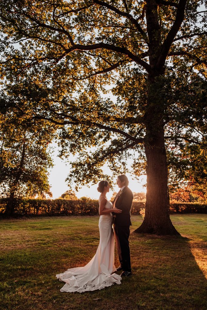 bride and groom stand under huge oak at sunset