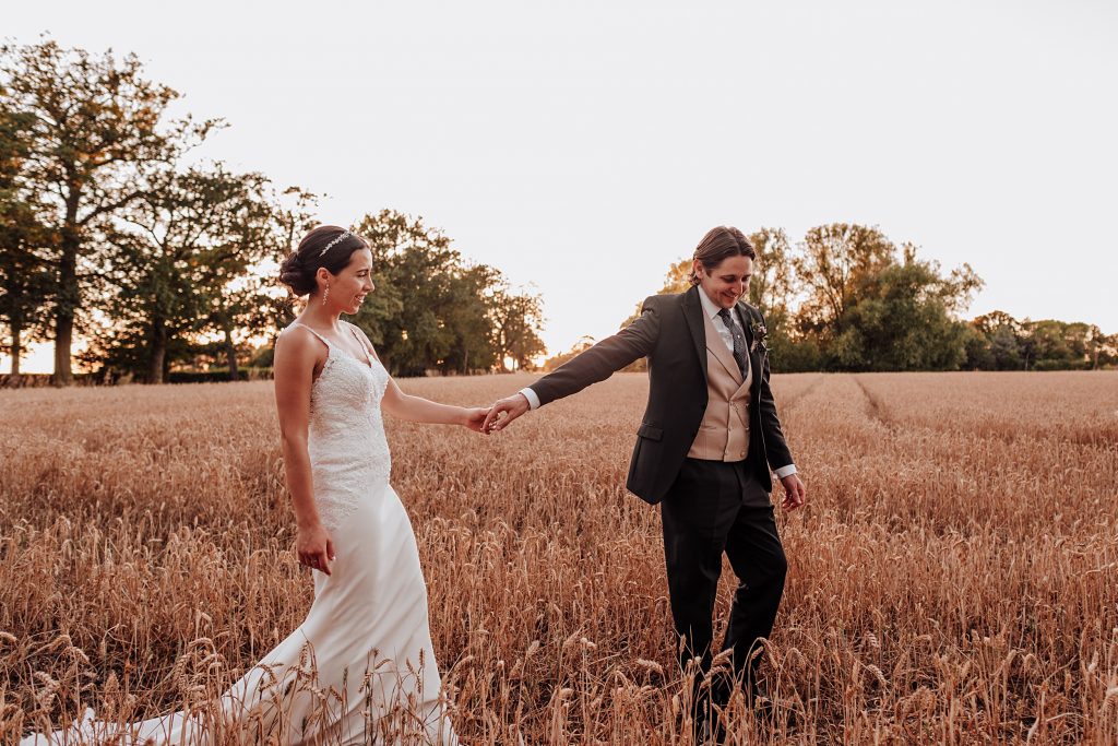 groom leads bride by hand as they walk into wheat field at sunset