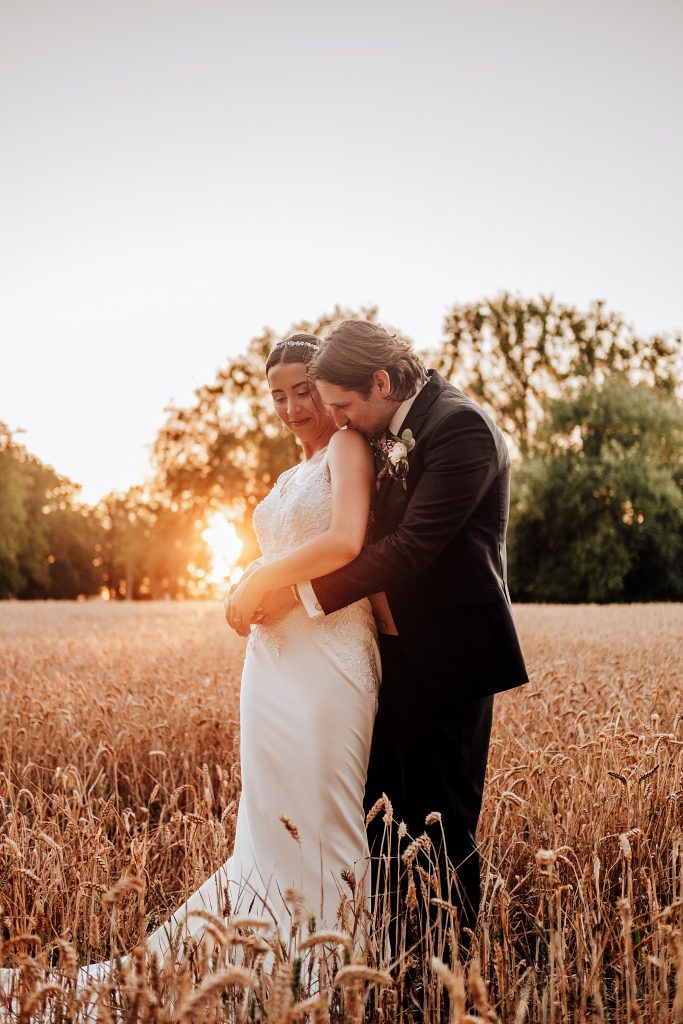 groom kisses brides shoulder in wheat field at sunset