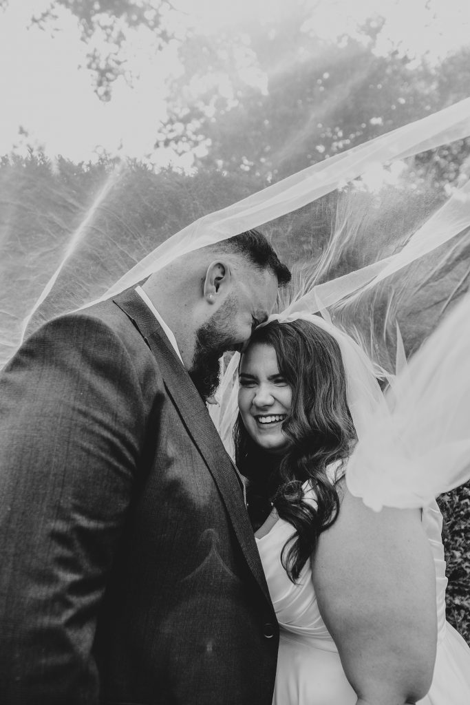bride and groom laugh under a billowing veil in garden