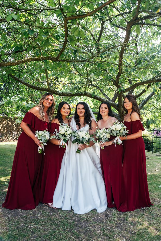 full length of bride with bridesmaids in red under a tree