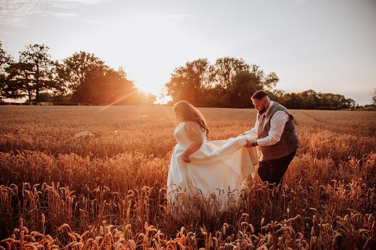 groom holds brides dress as they walk through wheat field at sunset