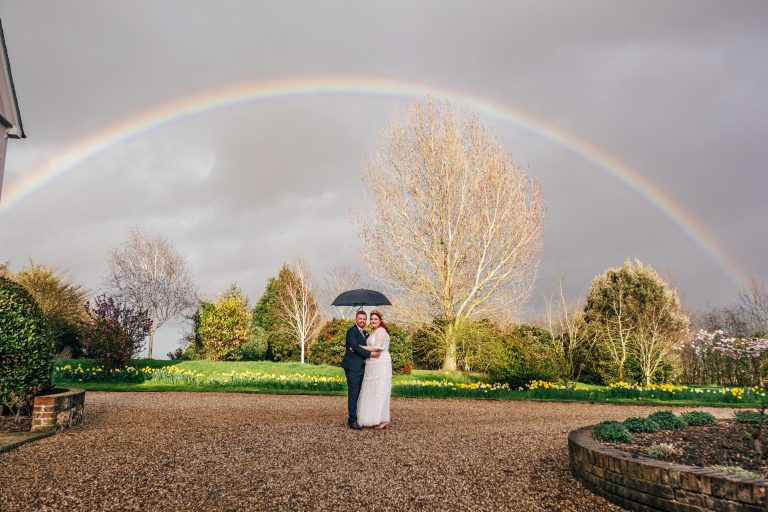 Bride and groom holding umbrella stand underneath a rainbow at Tuffon Hall
