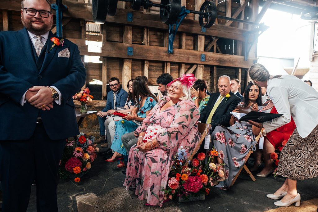 Grooms mum looks at him with pride as he waits for bride in rustic barn