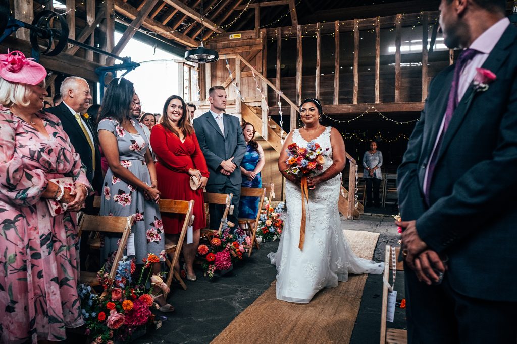 bride walks down aisle alone to meet groom in rustic barn