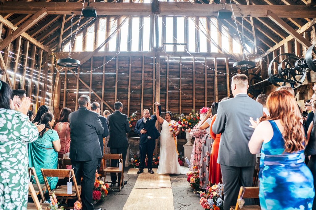 bride and groom raise hands in triumph as they prepare to walk up aisle in rustic barn