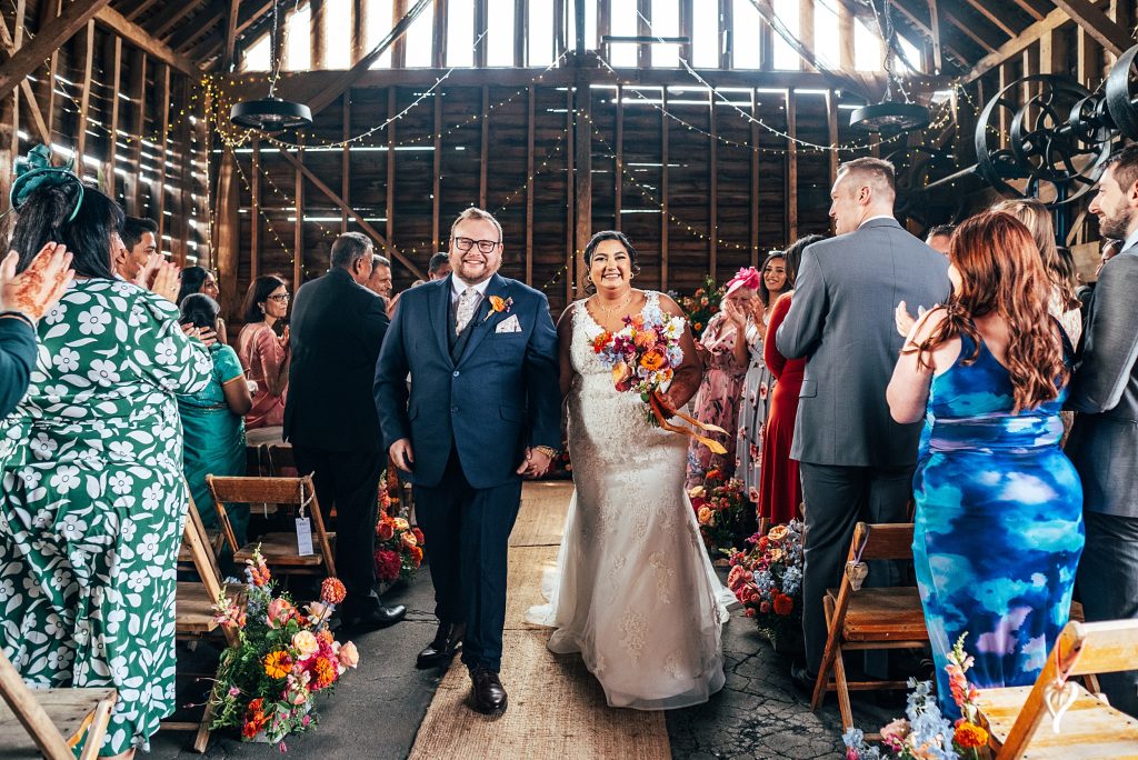 smiling bride and groom walk back up aisle in rustic barn