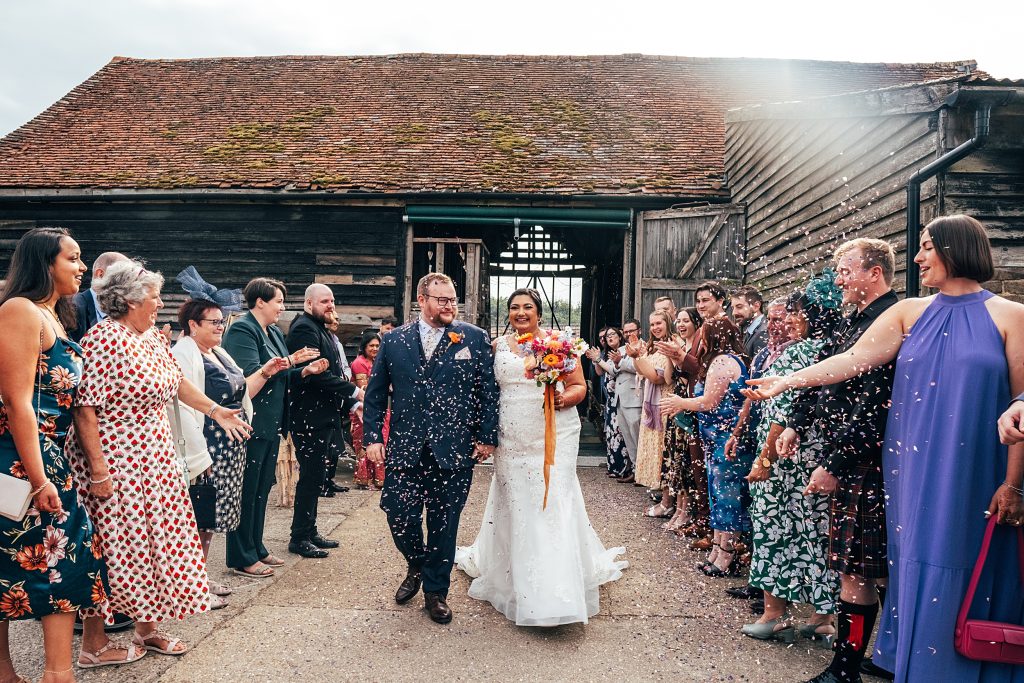 guests throw confetti on bride and groom as they emerge from barn