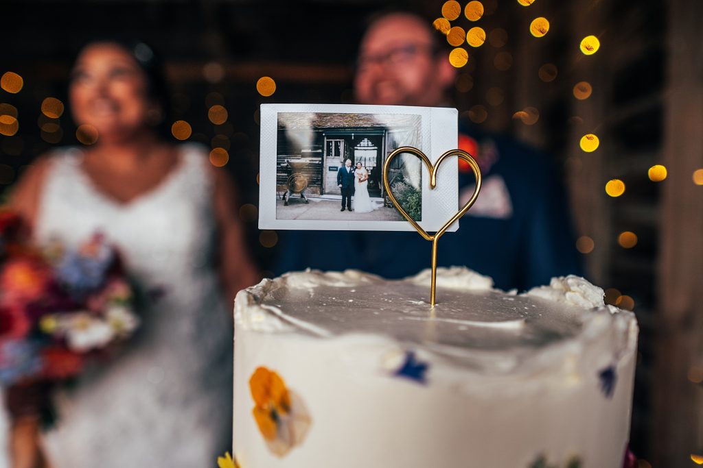 close up of cake topper - a gold wire heart holding a polaroid photo of the bride and groom with the couple in the background