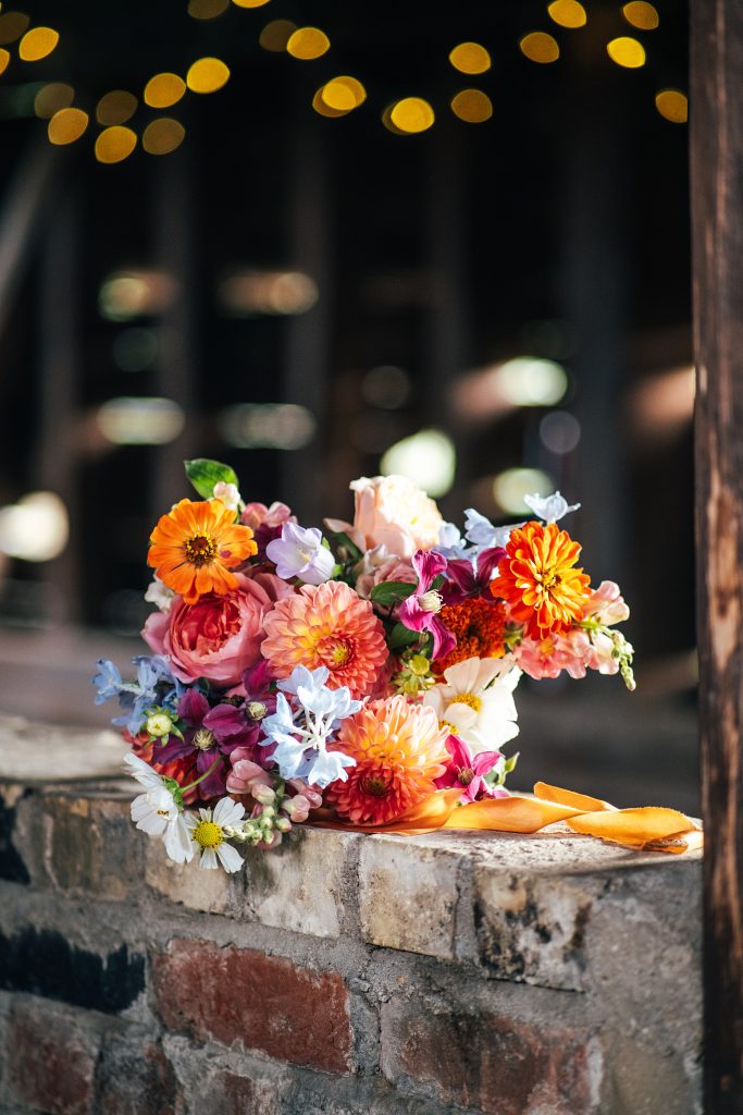 big, blousy, bouquet, oranges, pinks and lilacs with orange silk ribbon on wall in barn lit by fairy lights