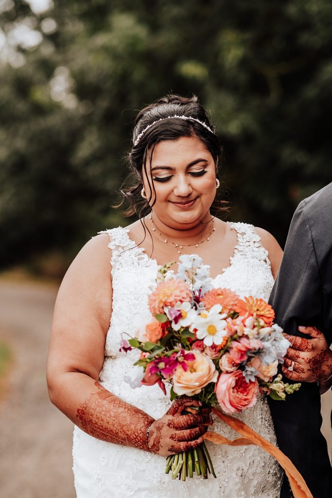 bride holds onto grooms arm out of frame and looks down at her orange and pink bouquet. Her arms and hands are decorated with henna