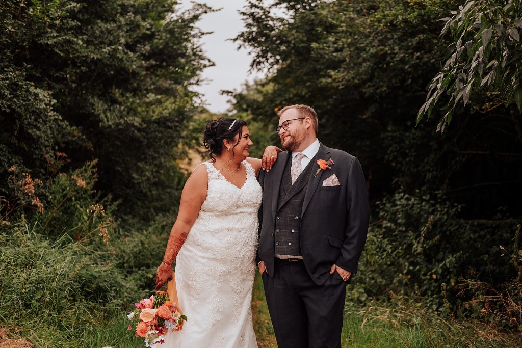 bride and groom smile at each other as she leans on his shoulder in a tree line