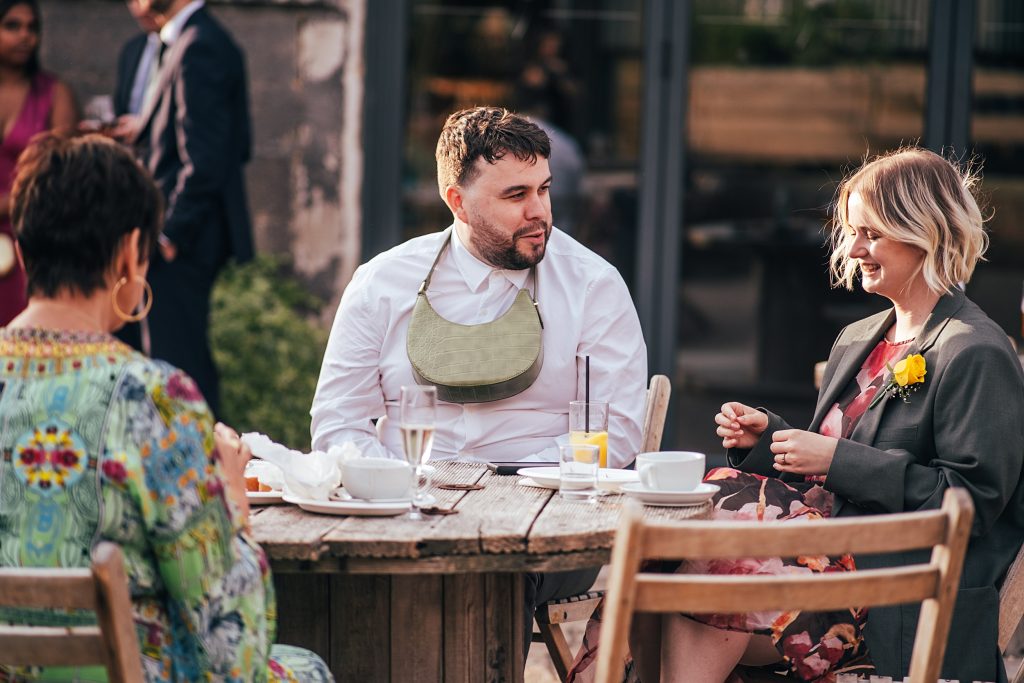 wedding guests chat around outdoor table. Man has handbag round his neck