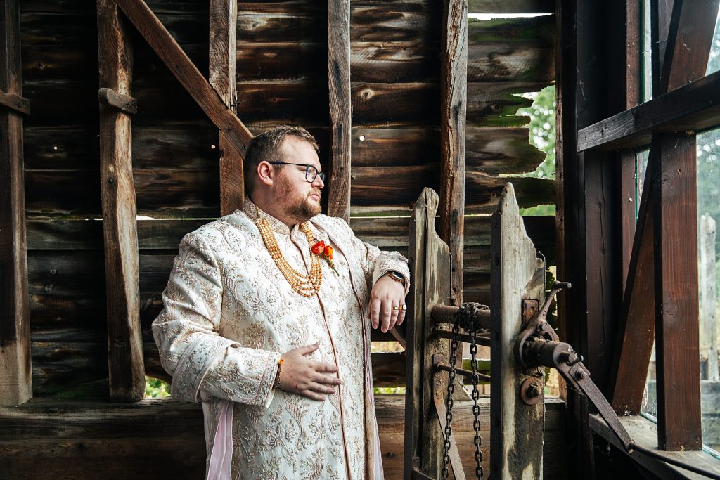 Groom in traditional Indian Sherwani looks out of window in rustic barn