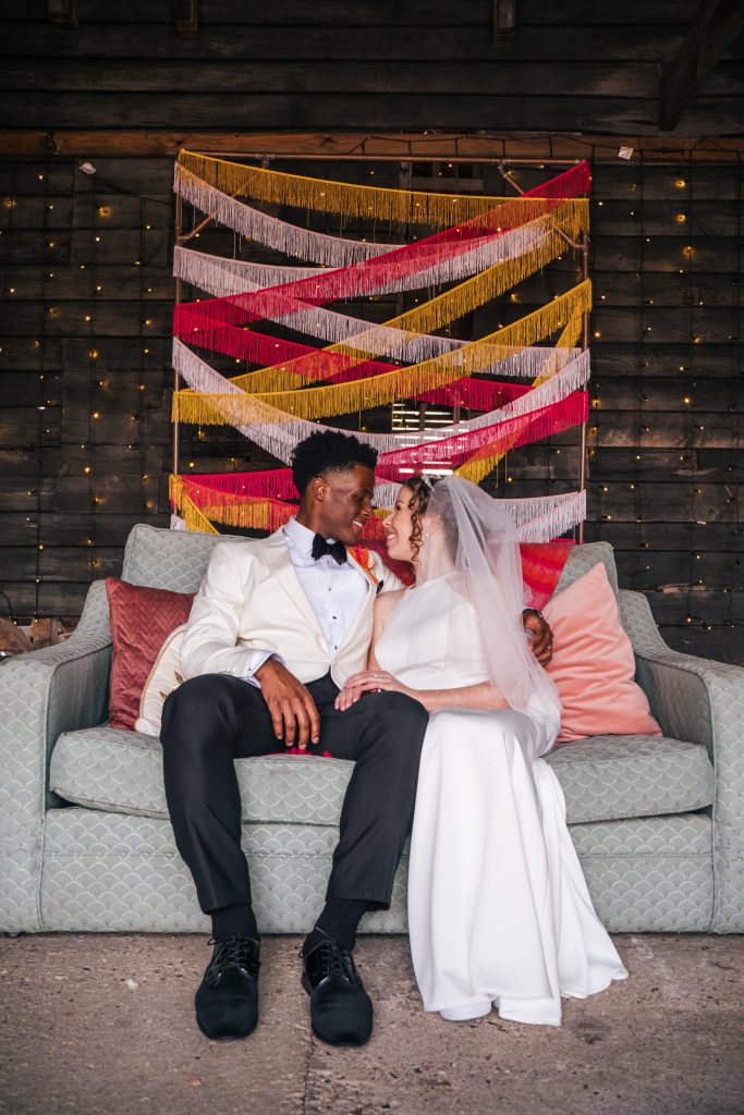 Groom in black tie with bride on blue sofa and pink and gold tassel wall hanging at The Barns at Lodge Farm Essex documentary wedding photographer