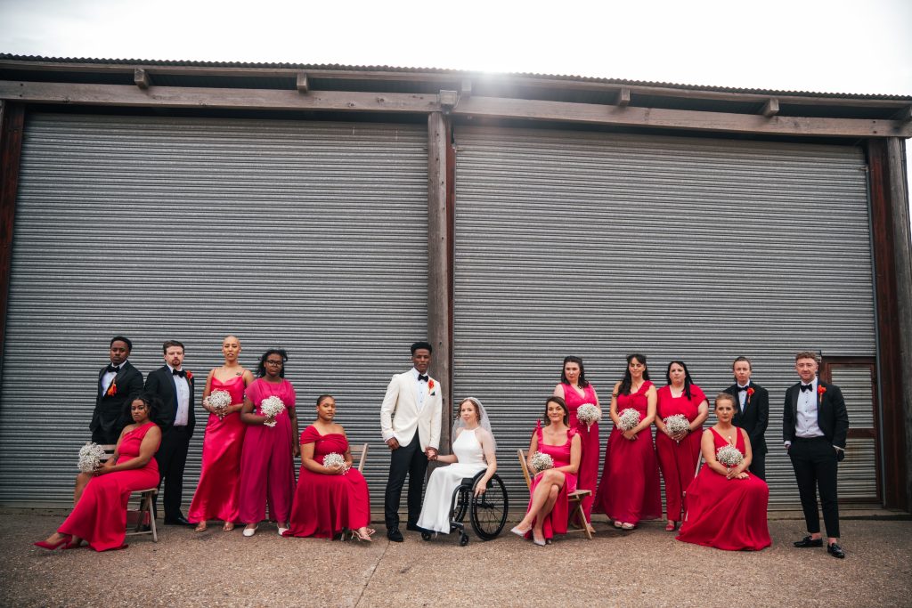 Bride and Groom with wedding party in black tie and hot pink stand outside corregated barn door Essex documentary wedding photographer
